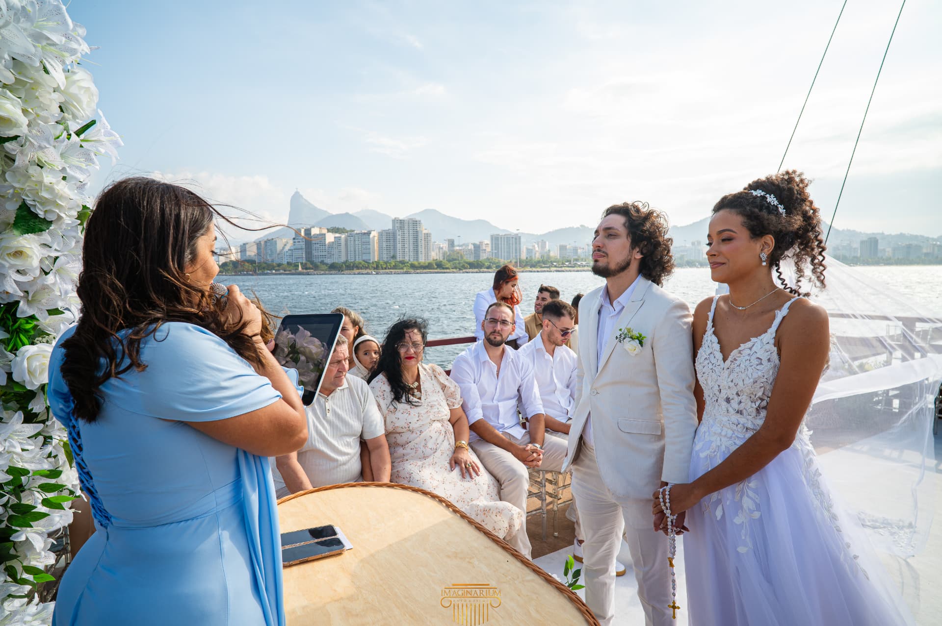 Celebrante de vestido azul conduz a cerimônia no convés enquanto os noivos se olham, com o Cristo Redentor e a orla ao fundo